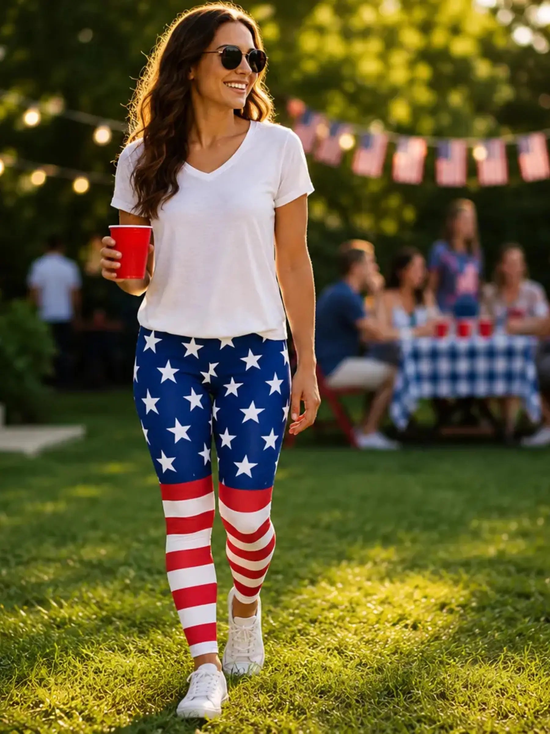Woman wearing American flag leggings and sunglasses, holding a red cup, in a park setting with people and flags in the background.