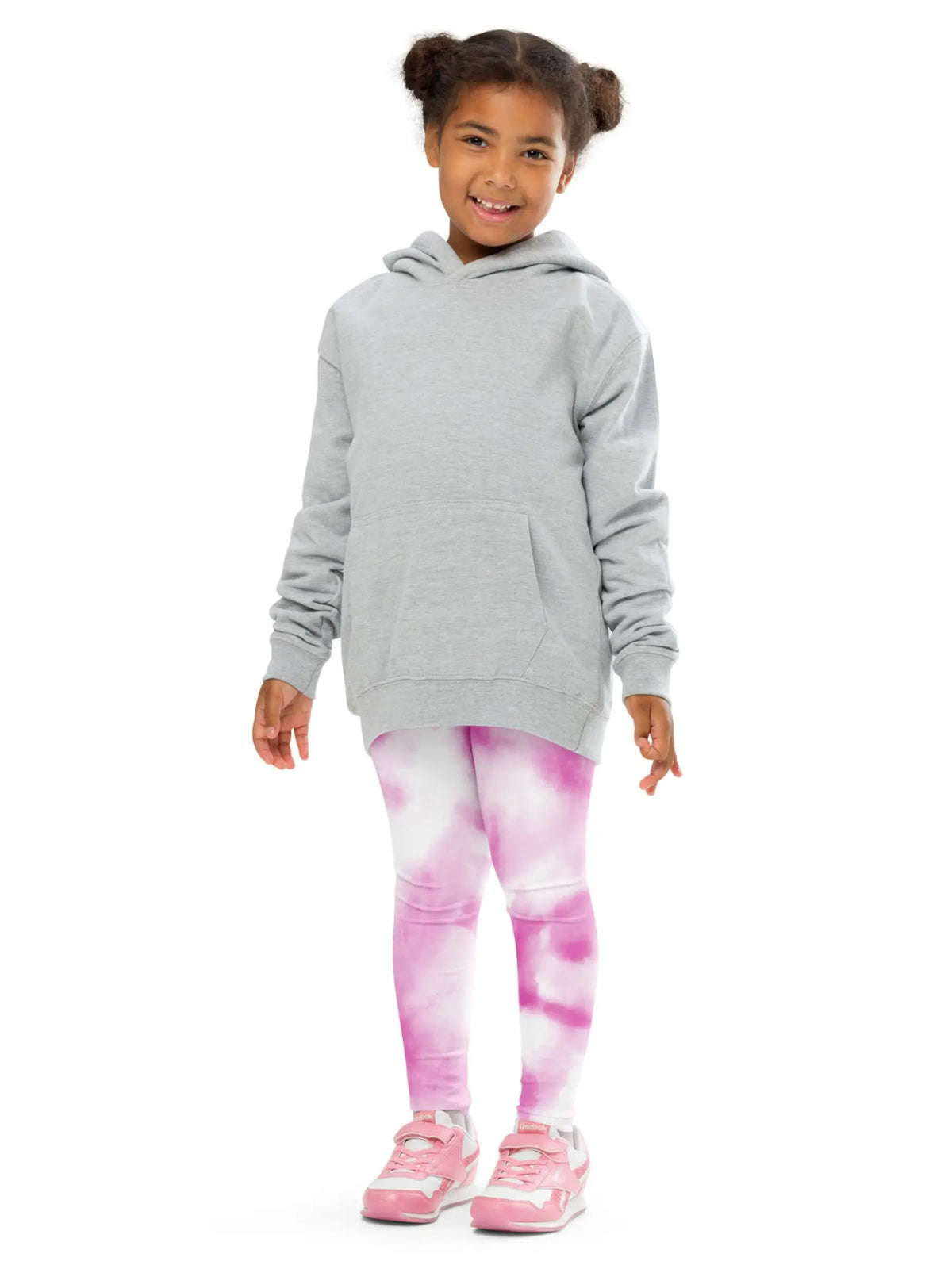 A young girl with her hair in two buns stands against a white background wearing a light gray oversized hoodie, pink and white tie-dye leggings, and pink sneakers.