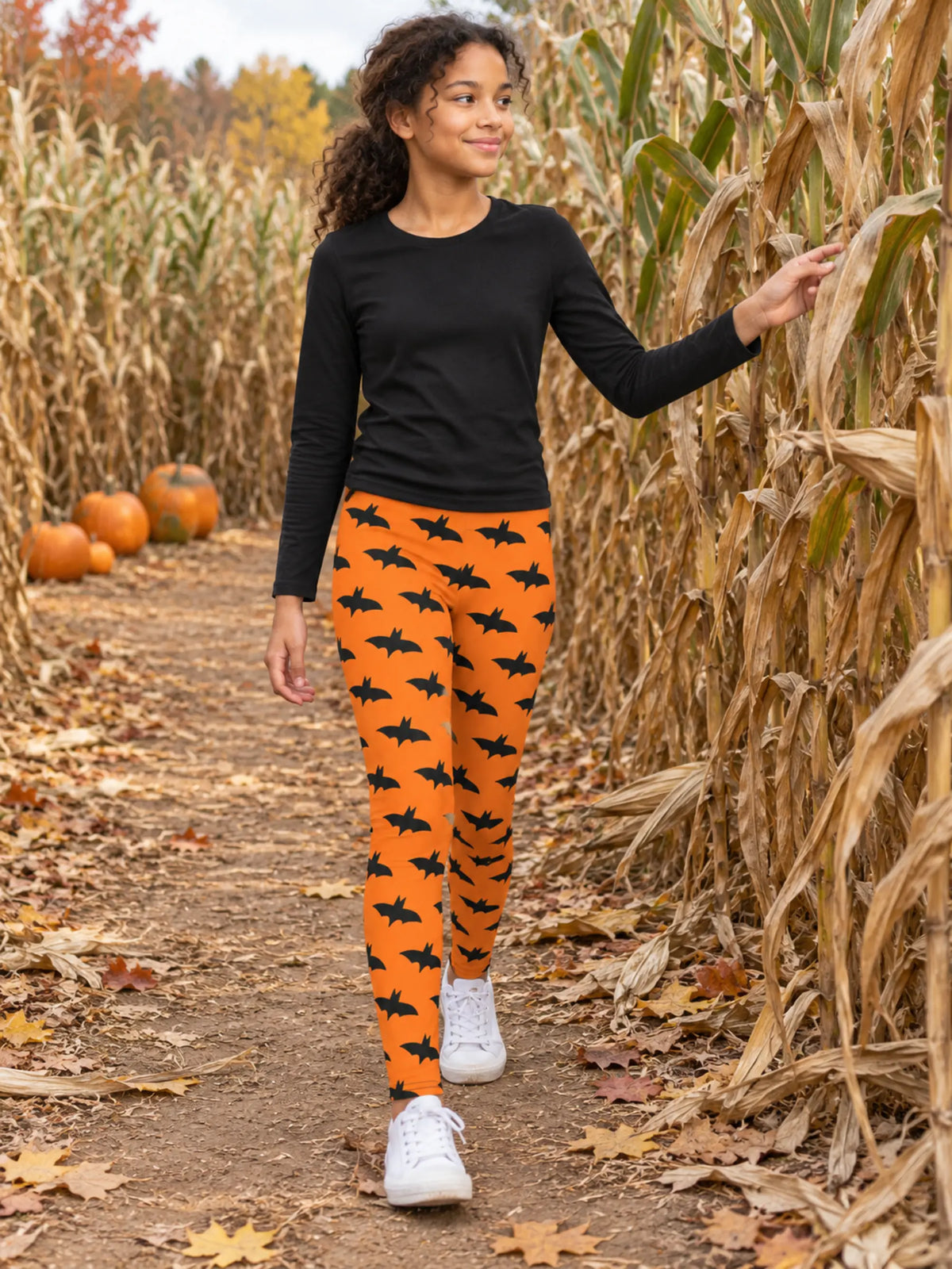 A woman walking through a pumpkin patch wearing bright orange leggings with a black bat pattern, a black long-sleeve shirt, and white sneakers.
