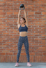 Load image into Gallery viewer, A person performing a yoga pose with a brick wall in the background. The person is wearing blue yoga leggings with a pattern and a black sports bra.