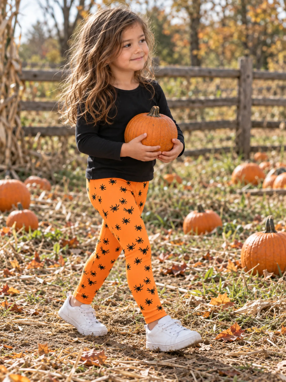 A young girl walking in a pumpkin patch wearing bright orange leggings with a black spider print, a black long-sleeve shirt, and white sneakers, while holding a small orange pumpkin.