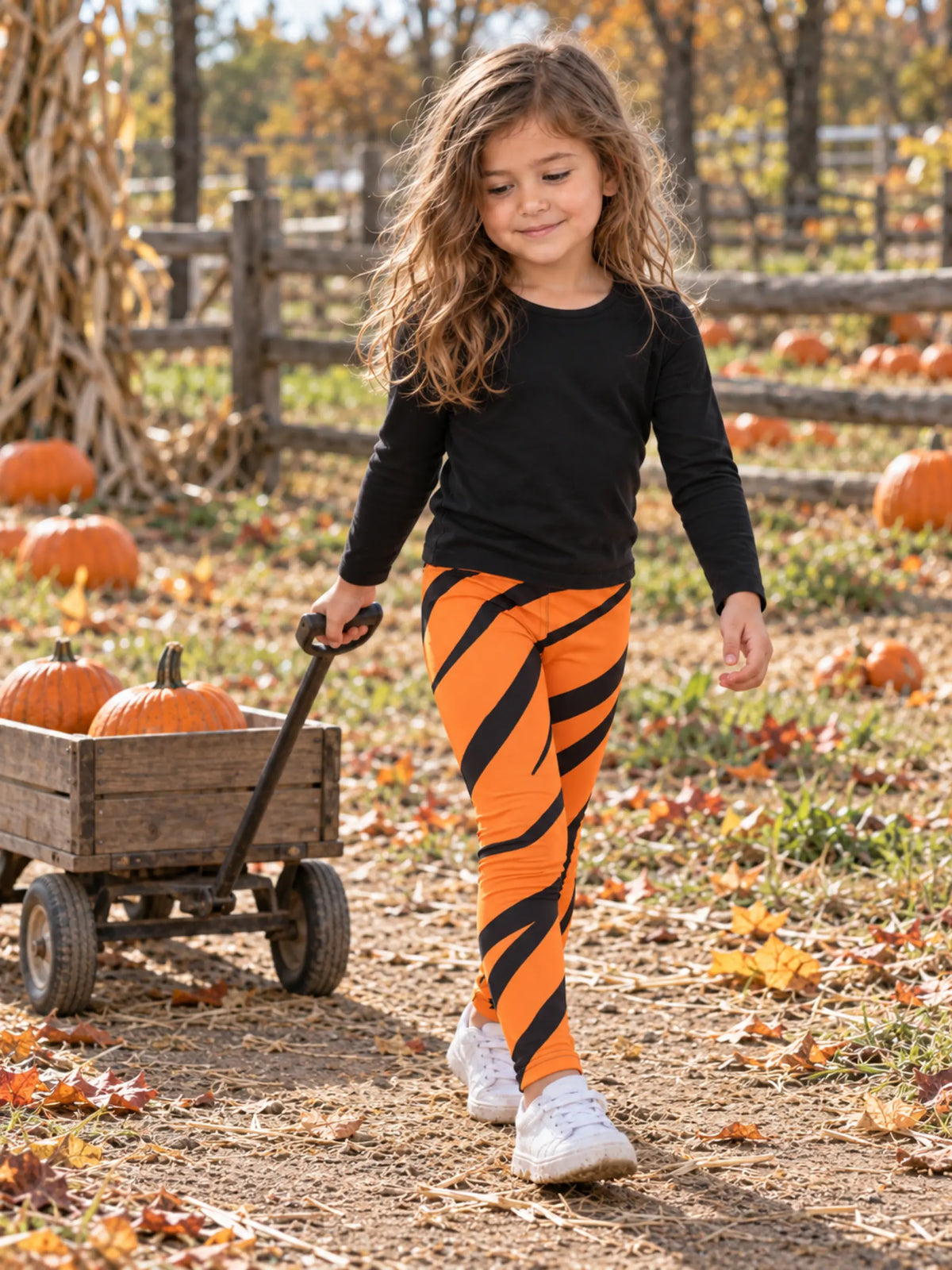 A young girl walking in an autumn pumpkin patch, wearing a black long-sleeve top and bright orange leggings with bold black diagonal stripes. She is pulling a small wooden wagon loaded with pumpkins.