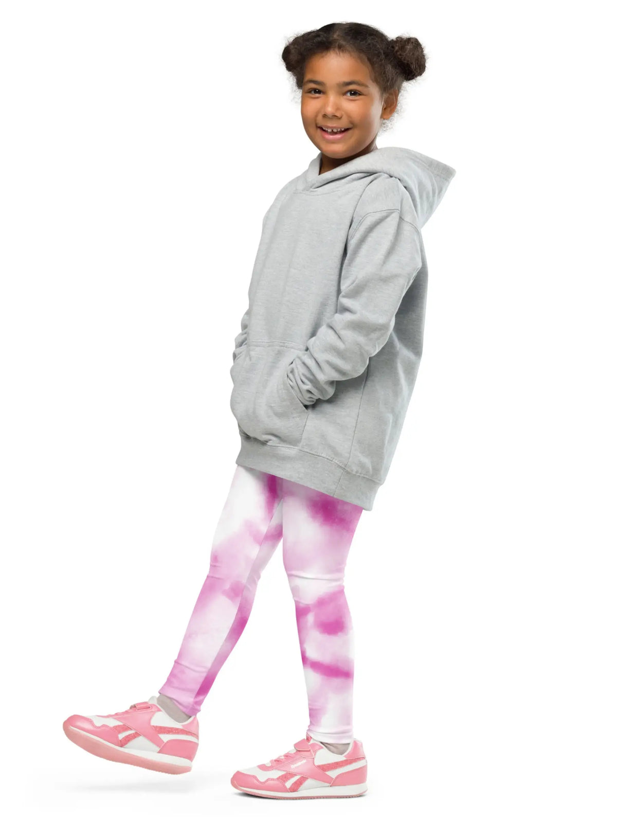 A young girl with her hair in two buns is shown from the side/back wearing a light gray oversized hoodie, pink and white tie-dye leggings, and pink sneakers against a solid white background.